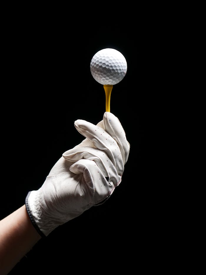 A gloved hand elegantly holds a golf ball on a tee against a black background.